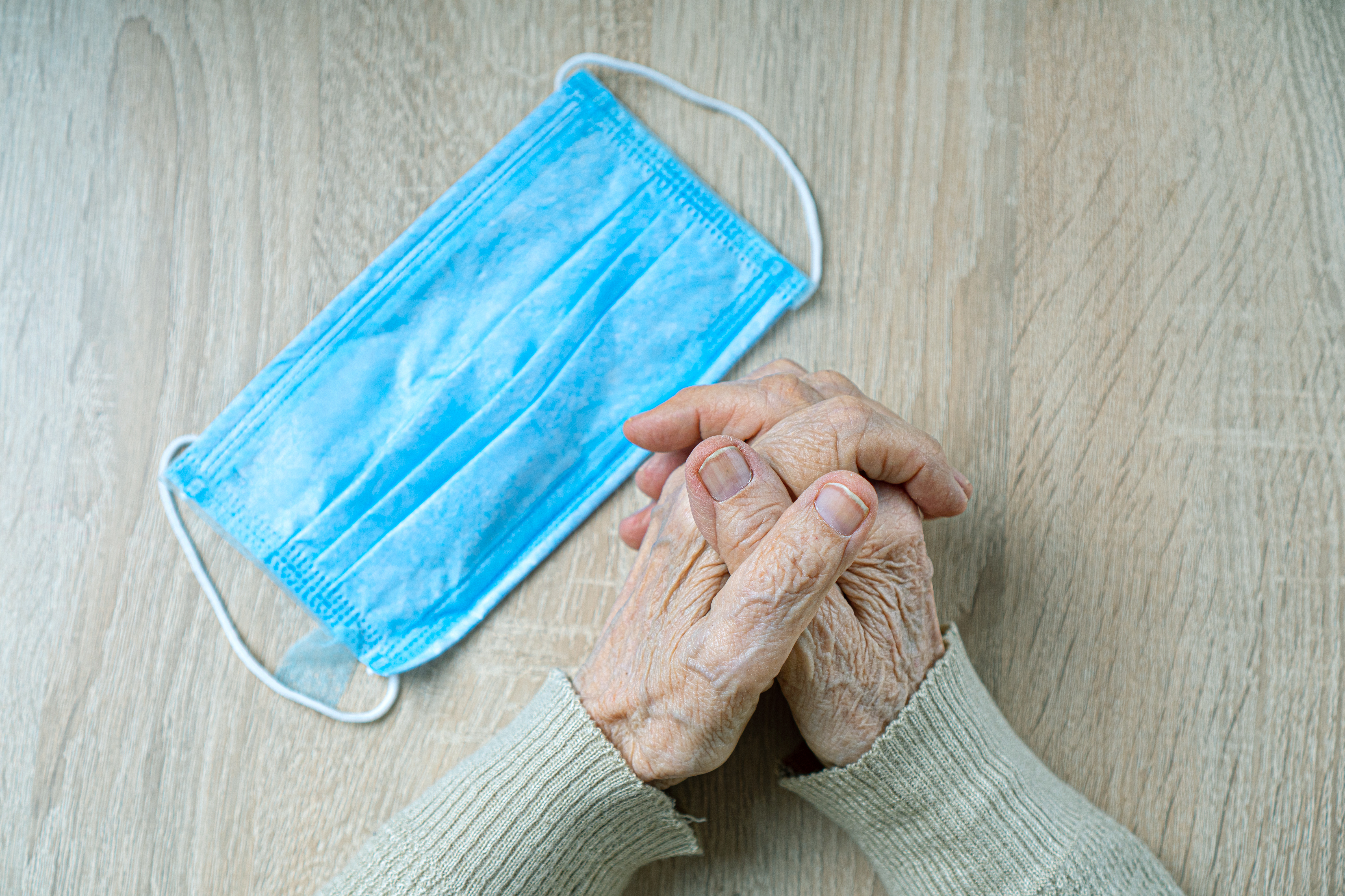 elderly hands resting on table with interlocked fingers. Surgical mask also on table.
