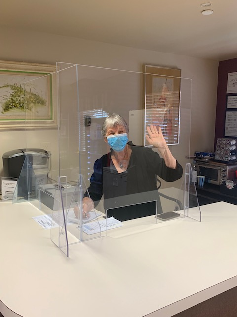 Woman standing behind counter, behind glass barrier, wearing surgical mask, waving happily